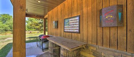 A covered outdoor patio with a wooden table and barrels, a grill, and a framed American flag.