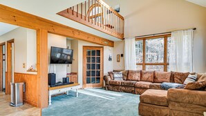 Ein geräumiges Wohnzimmer mit hohen Decken, einer Holztreppe, einem großen Fenster mit Vorhängen und einem braunen Ledersofa.