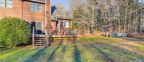 A two-story wooden house with a deck, surrounded by trees and a grassy yard.