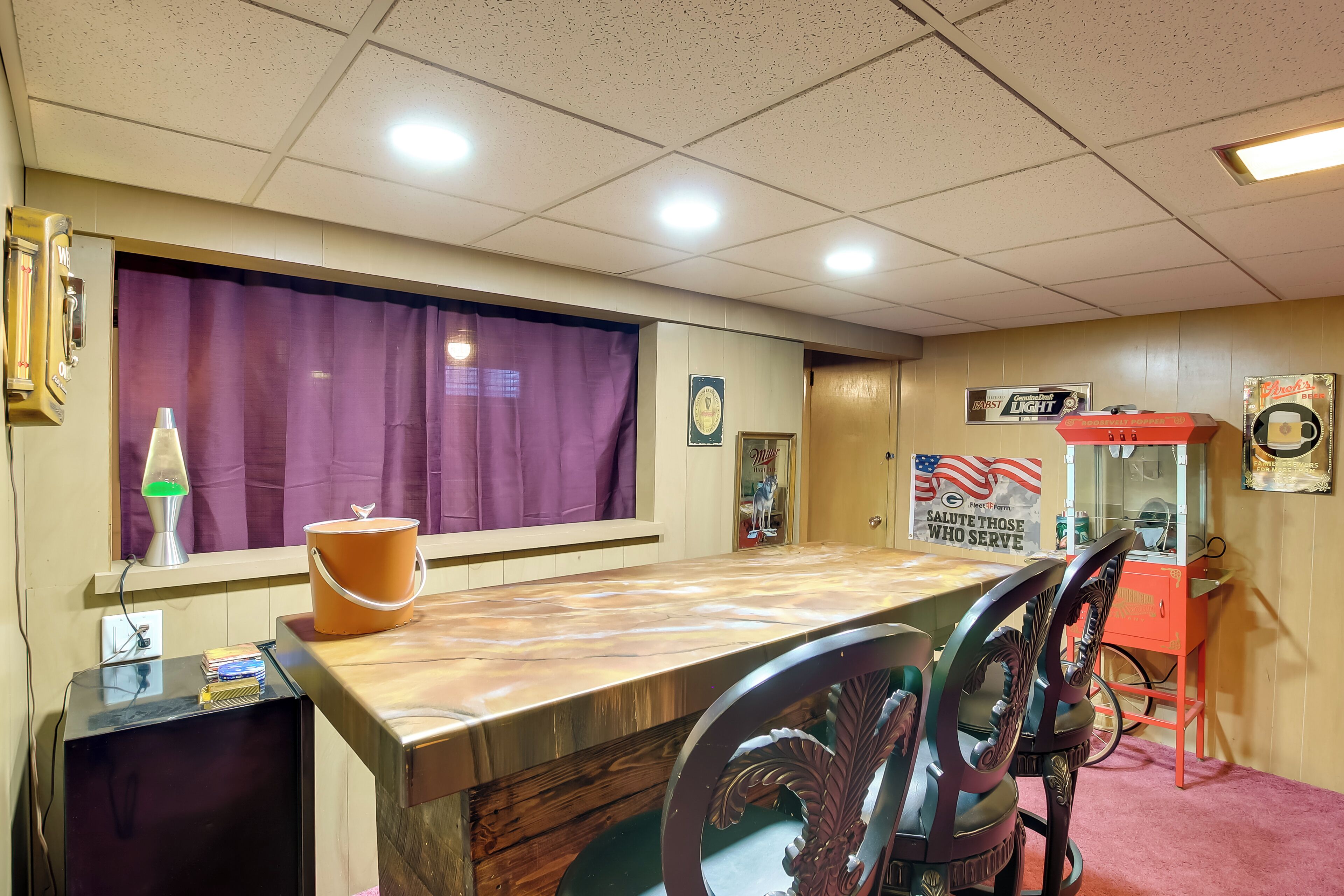 A room with a wooden table, purple curtains, a lava lamp, a bucket, a trash can, a vending machine, and a sign that says "Salute Those Who Serve".