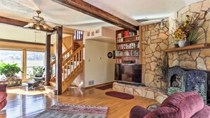 A spacious living room with a wooden staircase, a fireplace, a bookshelf, and a large TV. - South Dakota Vacation Rental Near Mount Rushmore (Hermosa)