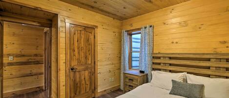 A wooden cabin bedroom with a bed, a nightstand, a window with curtains, and a wooden door.