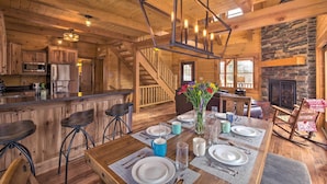 A wooden cabin interior with a dining area, kitchen, and a stone fireplace.