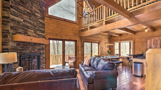 A spacious cabin interior with a stone fireplace, leather sofas, a dining area, and large windows offering views of the surrounding forest.