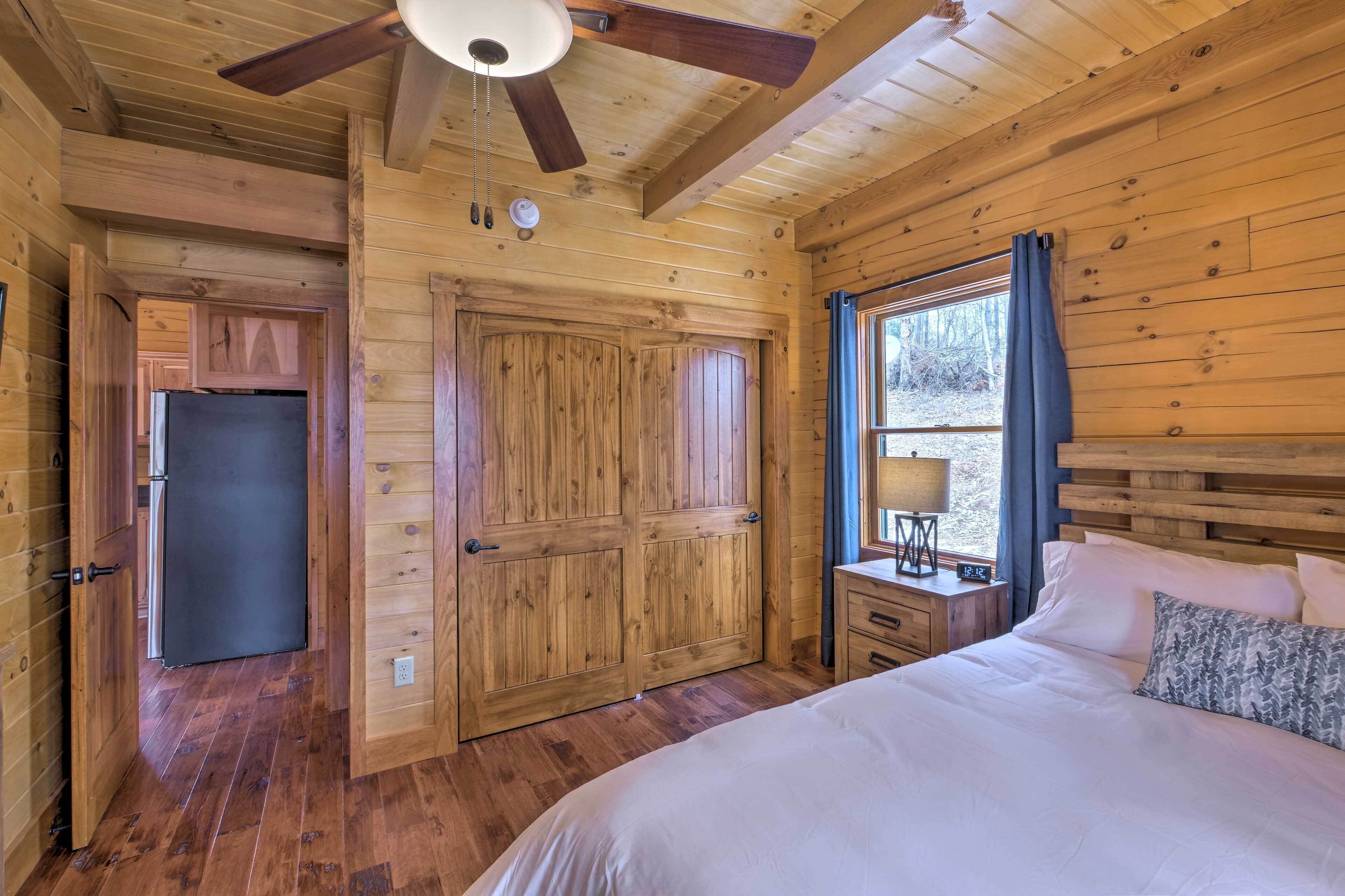 Un dormitorio de cabaña de madera con una cama, una mesita de noche, una ventana con cortinas y un ventilador de techo.