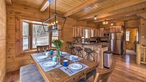 A wooden cabin with a dining area featuring a table set for four, a kitchen with modern appliances, and large windows offering a view of the surrounding forest.