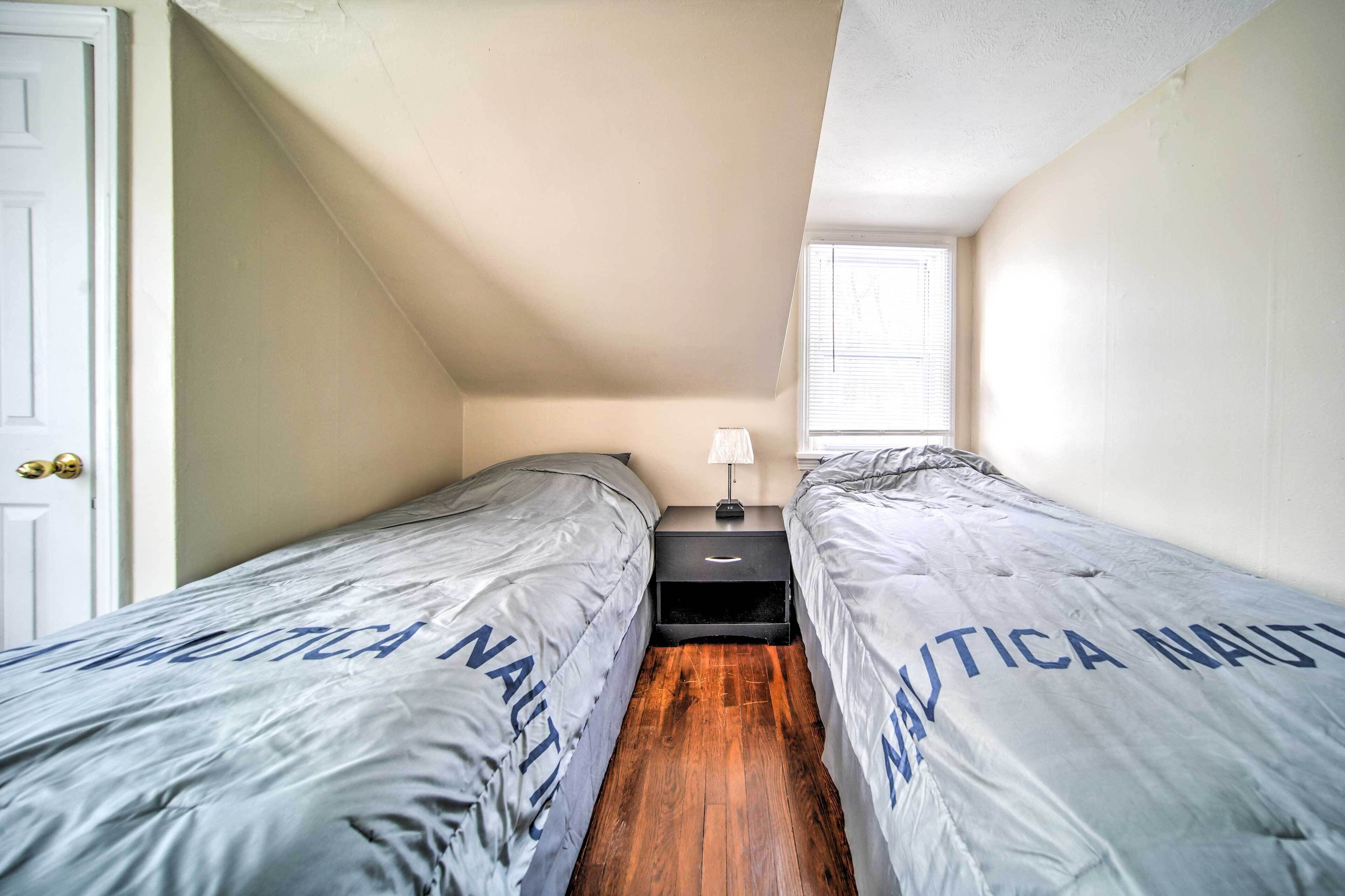 A small, narrow room with two beds, each covered with a grey blanket with 'NAUTICA NAUTICA' printed on it. There is a small nightstand with a lamp between the beds, and a window with blinds on the far wall.