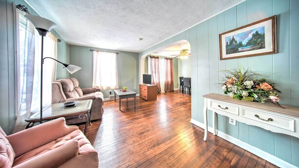 A living room with a wooden floor, a pink armchair, a sofa, a coffee table, a side table, a lamp, a dresser, a console table with a vase of flowers, and a framed picture on the wall. - Eastlake Home w/ Patio < 1 Mi to Lake Erie! (Eastlake)
