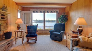 Un salón acogedor con dos sillones, una mesa de centro de madera, una chimenea y un amplio ventanal con vistas a un lago.