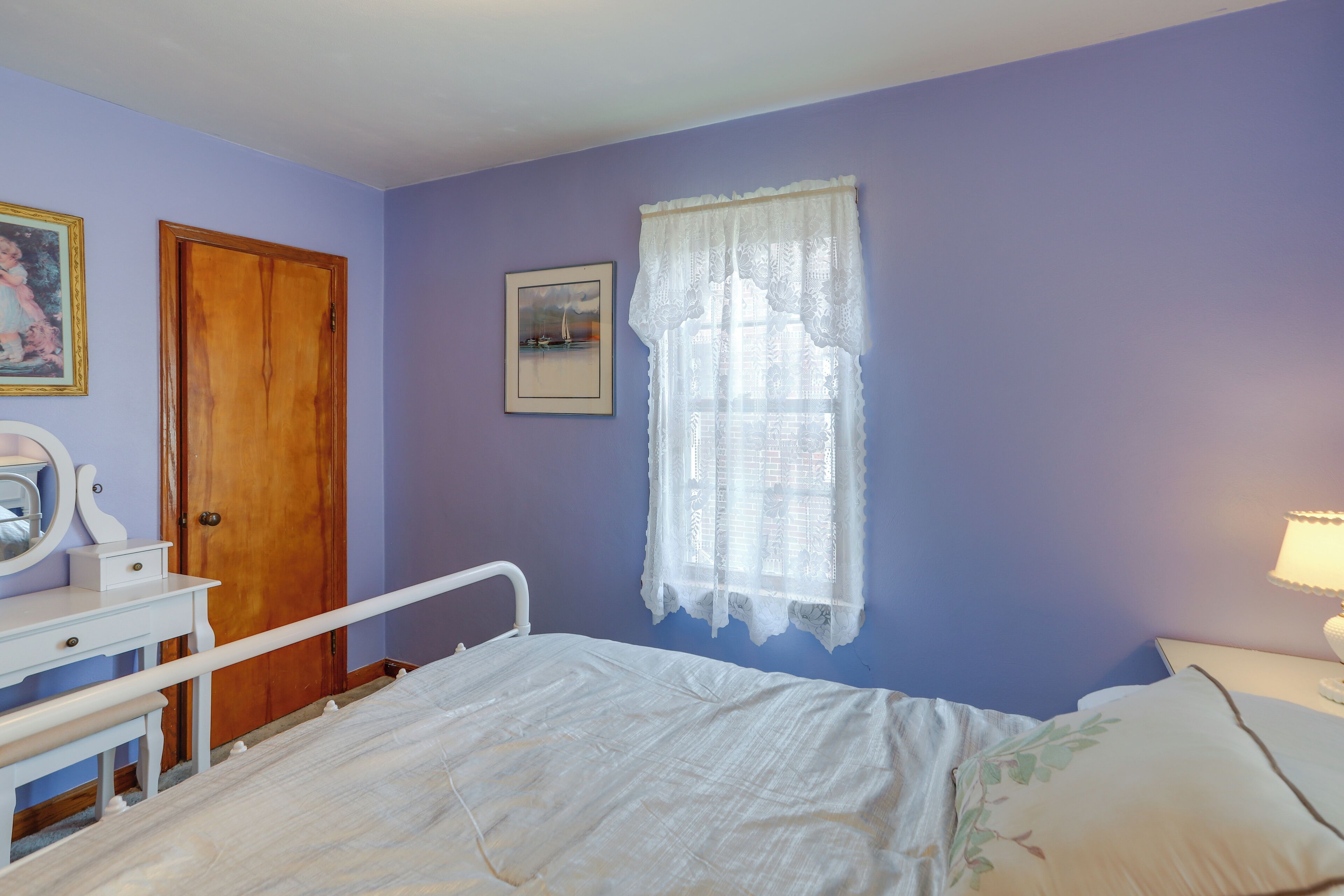Un dormitorio con cama, cómoda, espejo, puerta, ventana con cortinas de encaje y un cuadro en la pared.