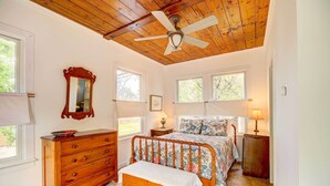 Une chambre avec un ventilateur de plafond en bois, un grand lit avec une literie à motifs floraux, une commode en bois et une fenêtre donnant sur des arbres.