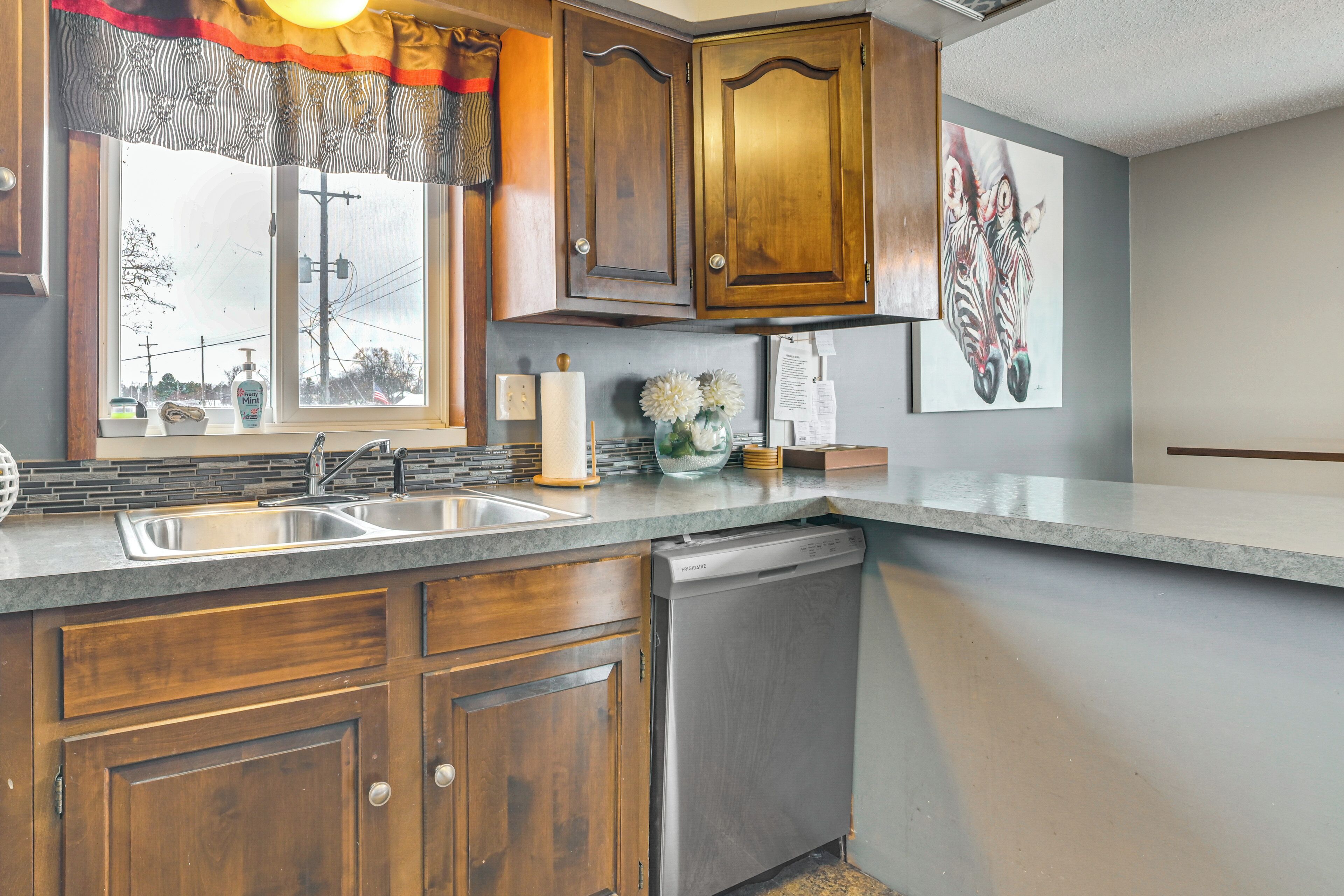 A kitchen with wooden cabinets, a stainless steel dishwasher, a double sink, and a window with a view of the outside.