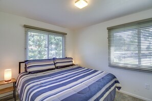 A bedroom with a bed, two windows with blinds, a bedside lamp, and a nightstand.