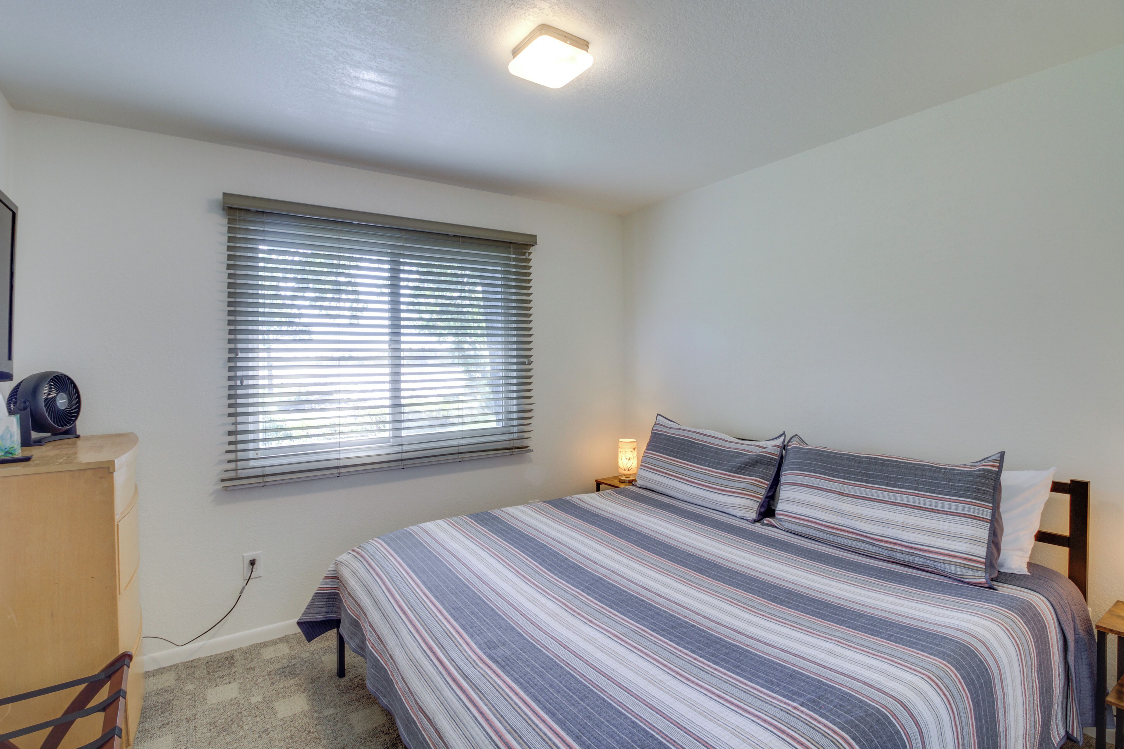 A bedroom with a bed, a bedside table, a lamp, a window with blinds, and a small desk with a fan.