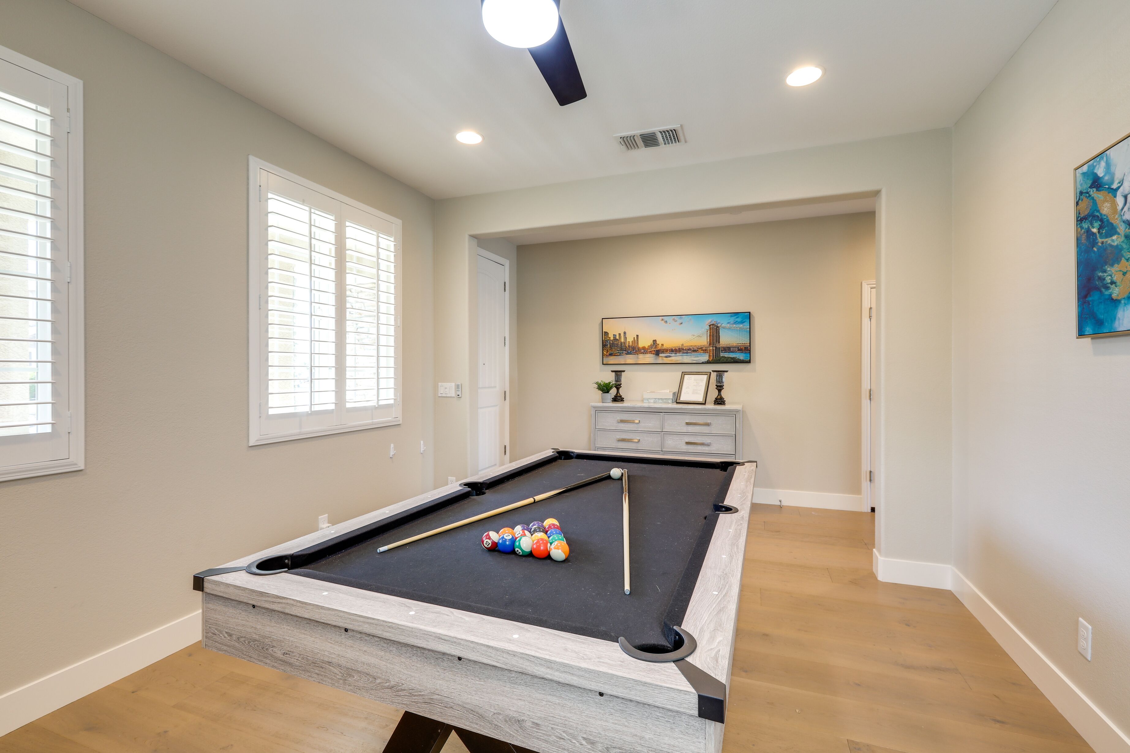 A pool table with balls, a painting, and a cabinet in a room with wooden flooring and white walls.