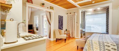 A bedroom with a wooden ceiling, a bed with a patterned bedspread, a chair, a small table, and a window with patterned curtains.
