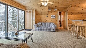 A cozy cabin interior with a wooden ceiling, a striped sofa, a glass coffee table, and a bar area with stools. - Lakefront Retreat w/ Patio, Grill + Boat Dock! (Eagle River)