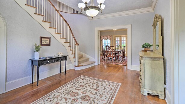 A spacious foyer with a wooden staircase, a chandelier, a dining area in the background, and a console table with a mirror.