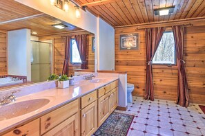 A wooden cabin bathroom with a large mirror, two sinks, a toilet, and a window with curtains.