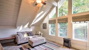 A cozy attic room with a sloped ceiling, a ceiling fan, a fireplace, and large windows offering a view of the trees outside. - Gilford Cabin - 2 Mi to Gunstock Mountain Resort! (Gilford)