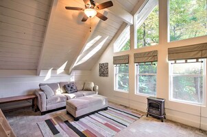 Un acogedor dormitorio en ático con techo inclinado, ventilador de techo, chimenea y ventanas amplias que dan a las copas de los árboles.