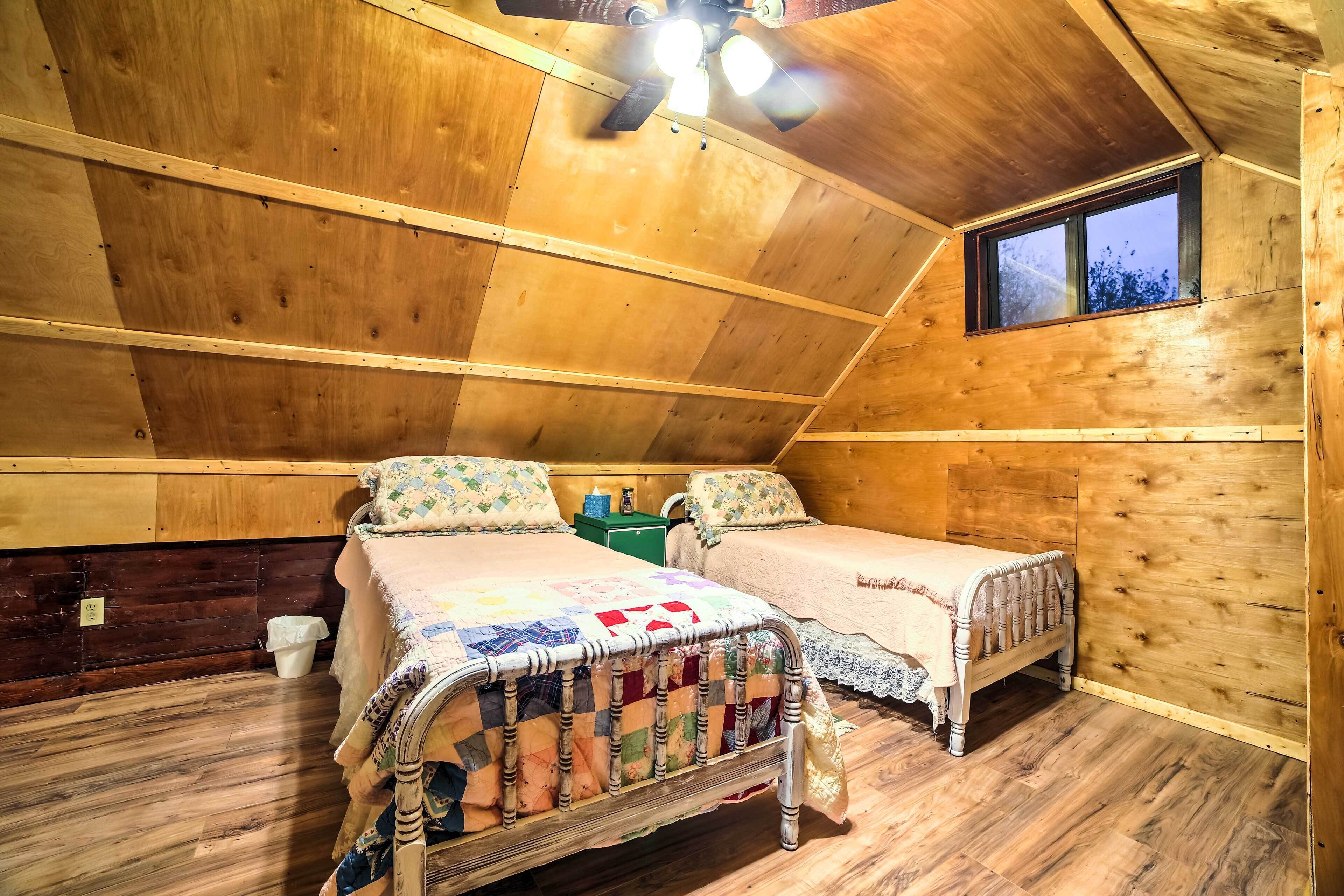 A cozy attic bedroom with two beds, wooden walls, a small window, and a ceiling fan.