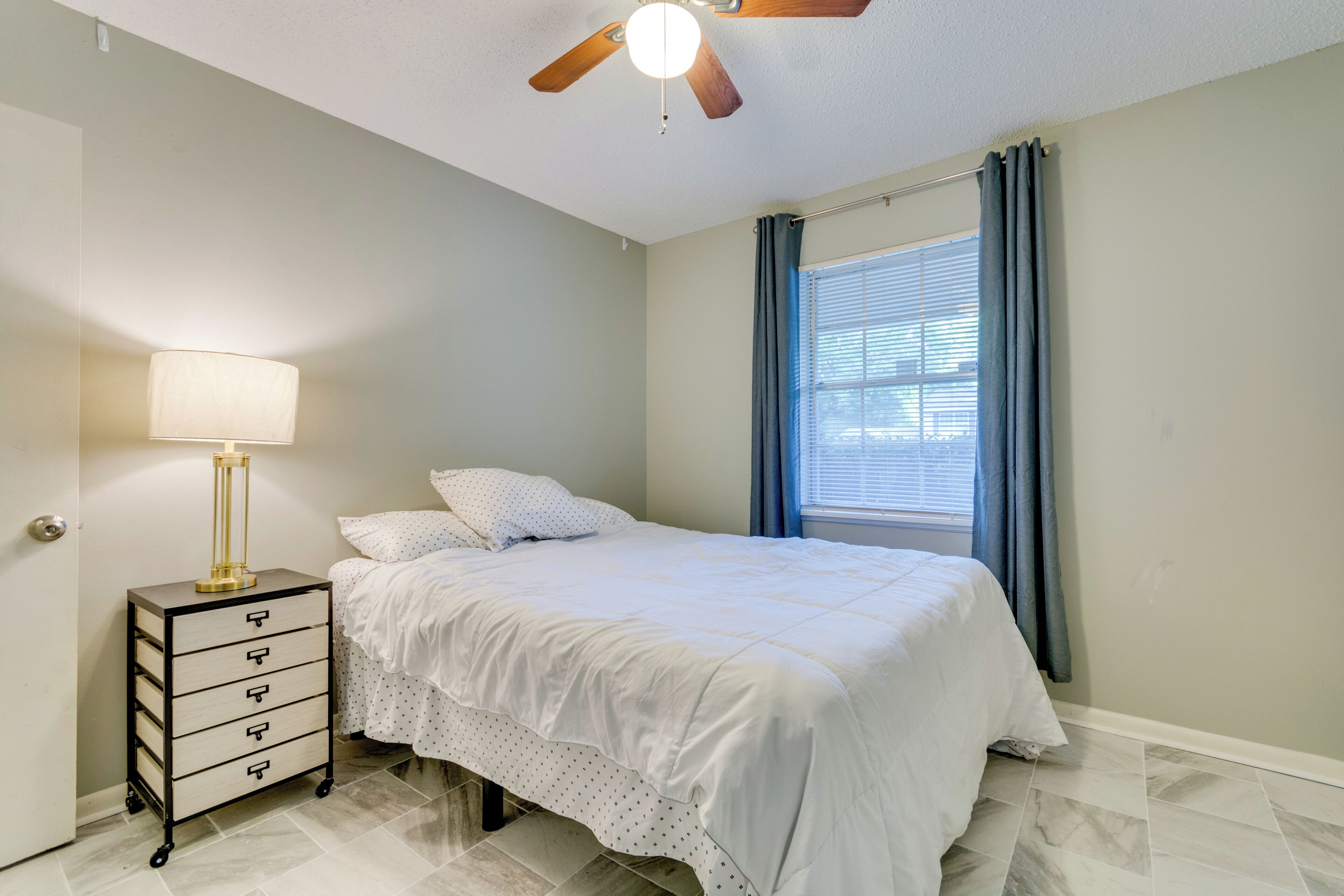 Une chambre à coucher avec un lit, une table de chevet avec une lampe, un ventilateur de plafond et une fenêtre avec des rideaux.