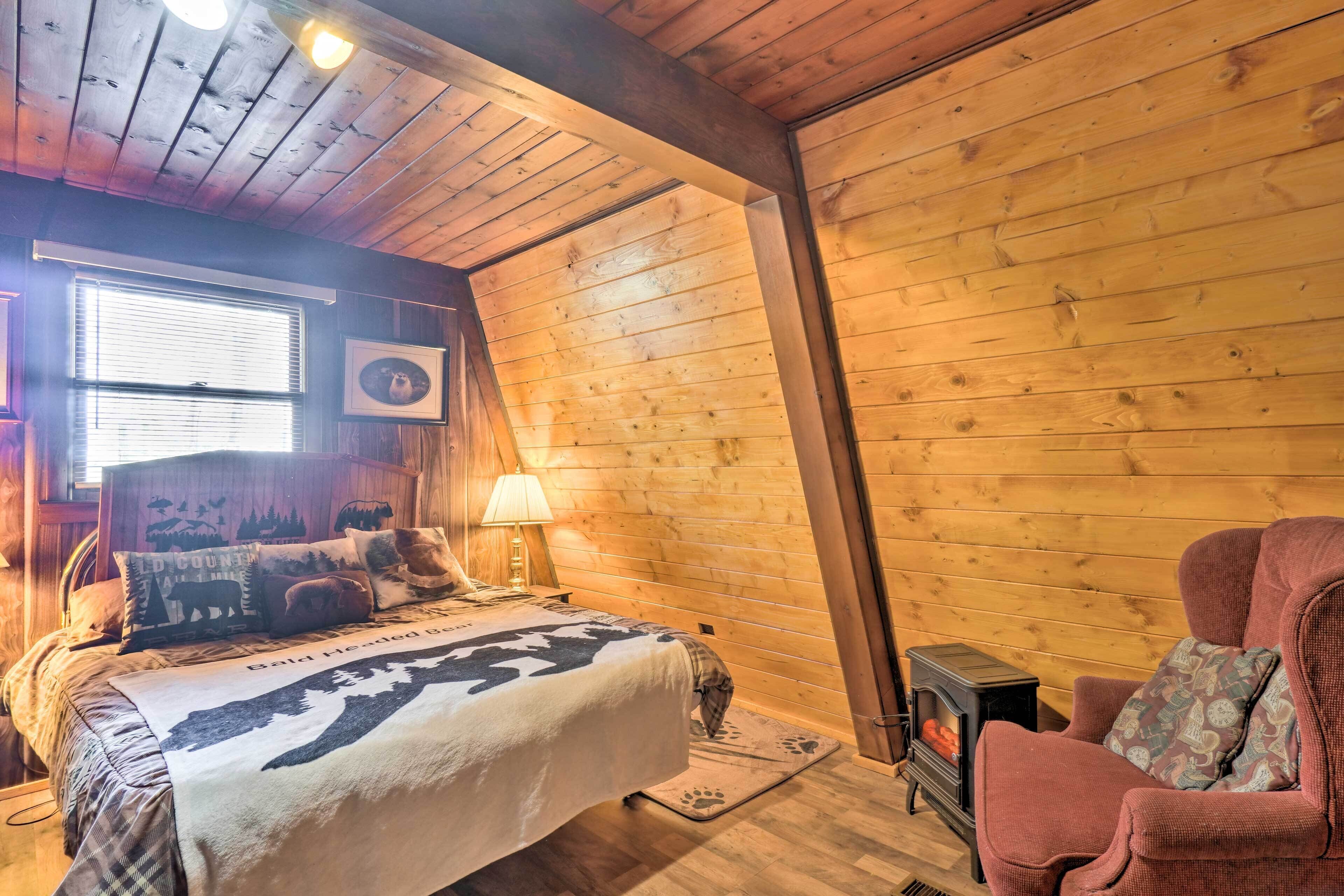 A cozy cabin room with a wooden ceiling, a bed with a blanket, a chair, a small heater, and a window with blinds.