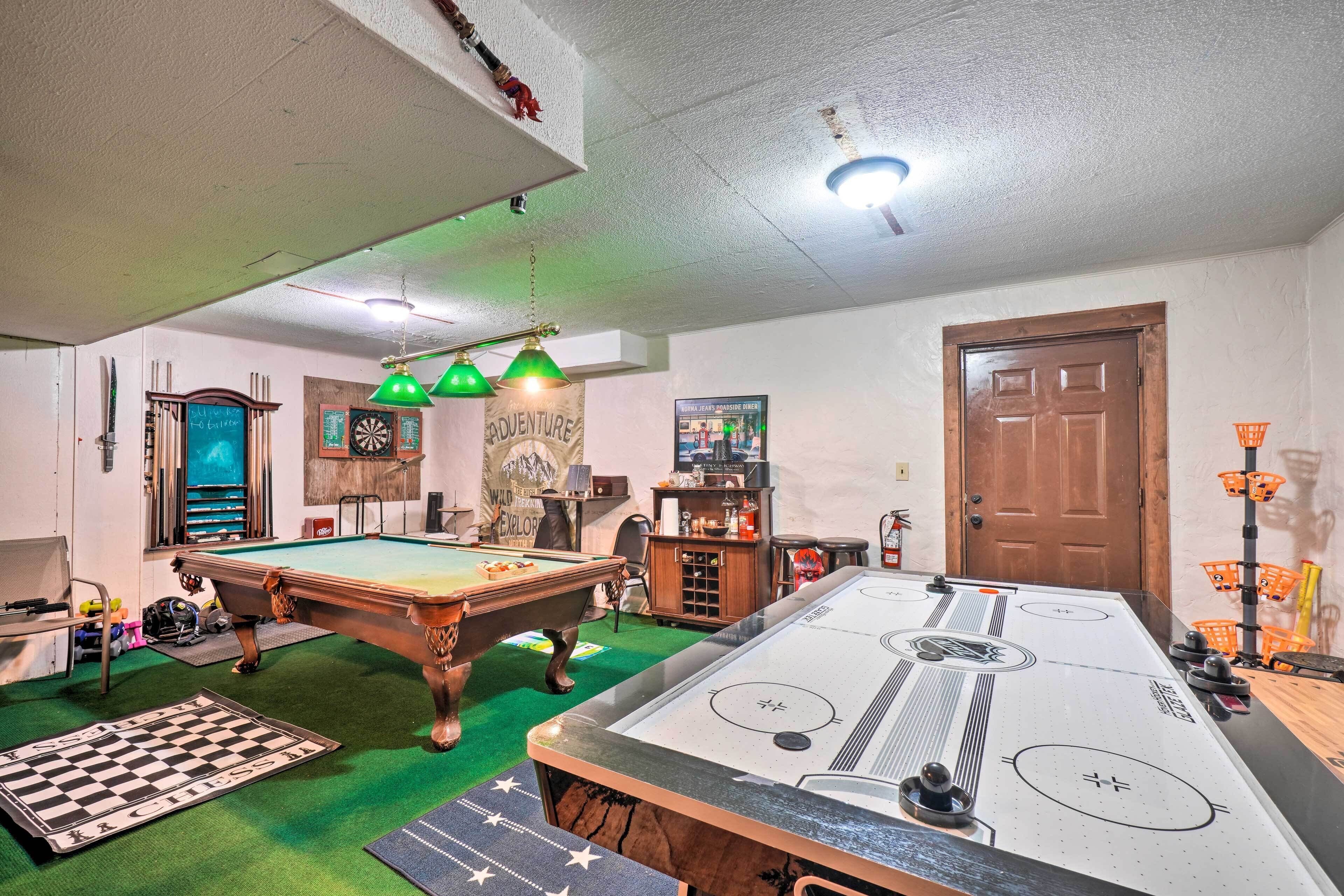 A pool table with a checkered mat, a foosball table, and a dartboard in a room with a green carpet and wooden cabinets.