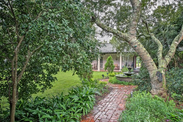 Um caminho no jardim leva a uma casa com uma fonte, cercada por vegetação exuberante e um gramado bem cuidado.