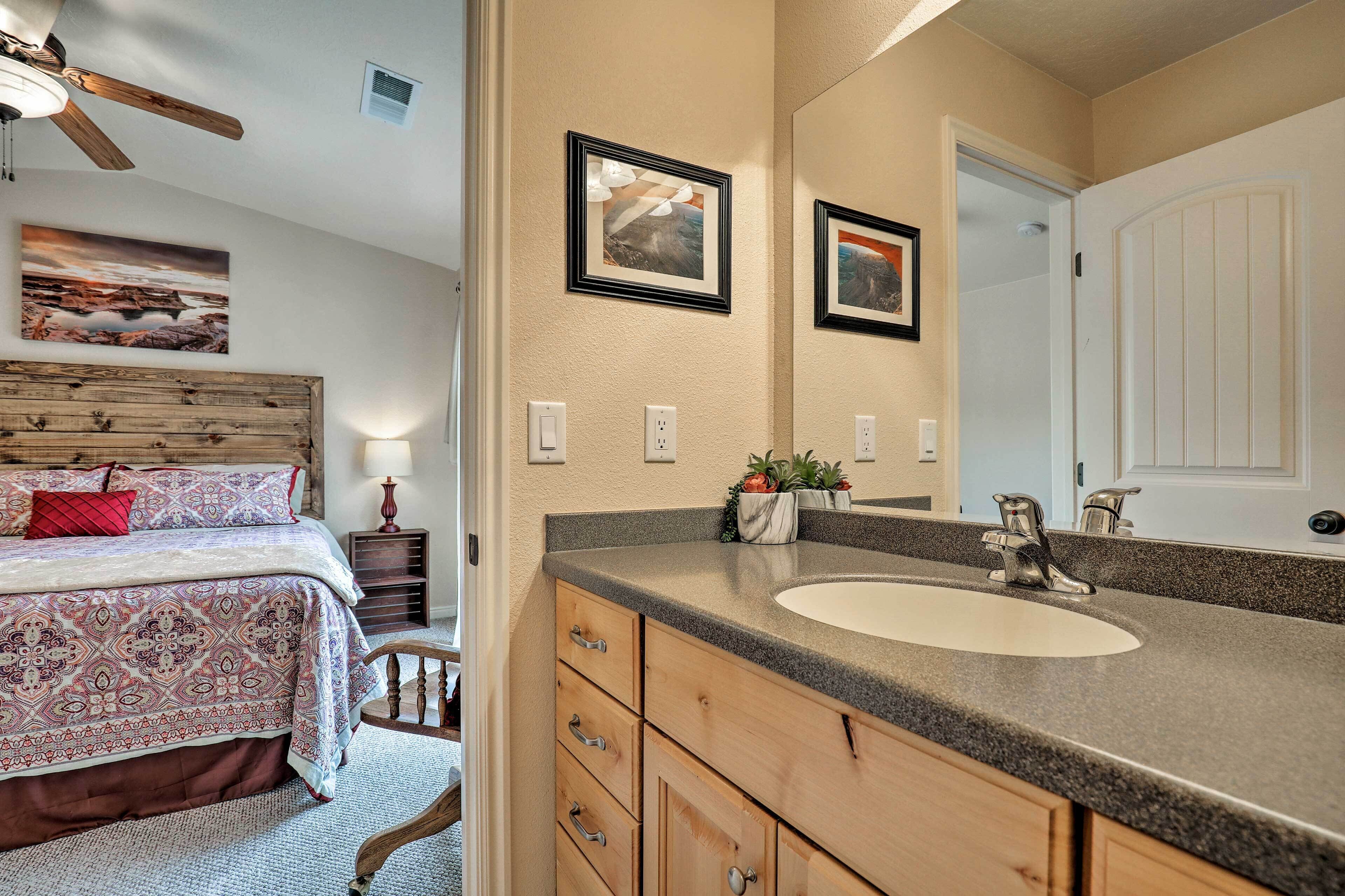 A bedroom with a bed, a wooden headboard, a nightstand, a mirror, and a bathroom with a sink and cabinets.