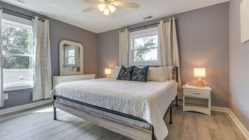 Quarto com cama, mesas de cabeceira, ventoinha no teto, janela com cortinas e espelho na parede.