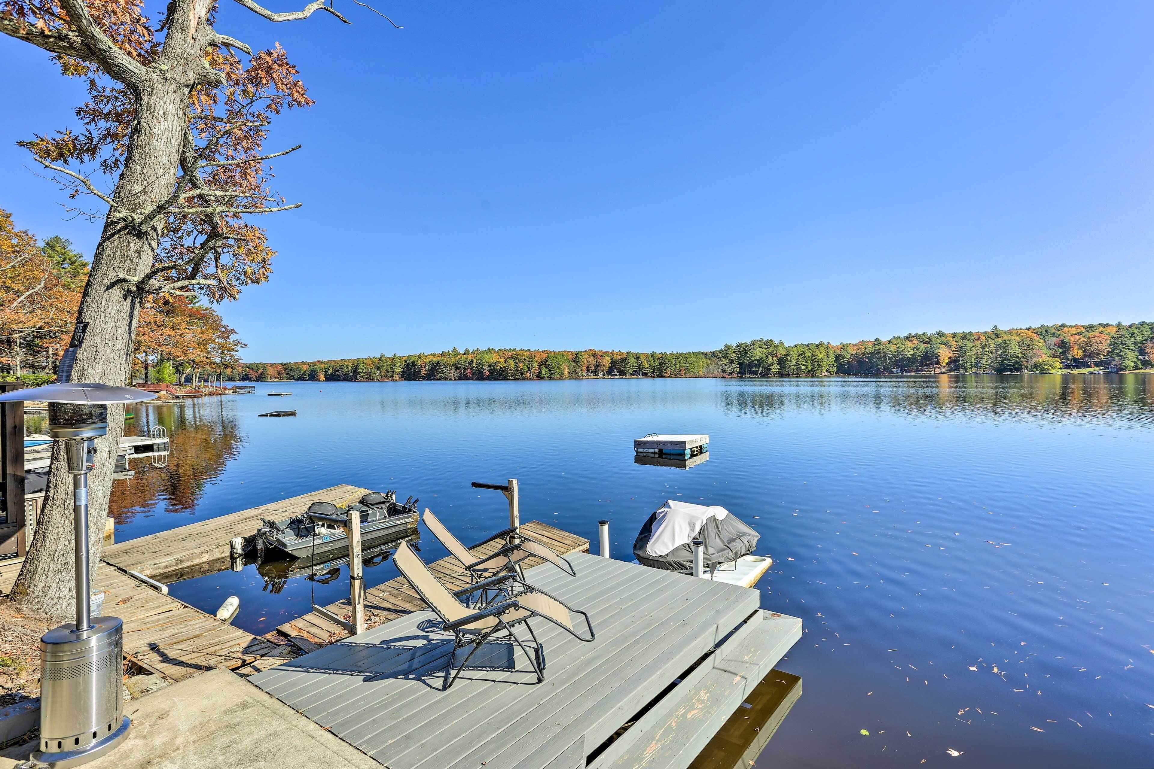 A dock with chairs and a grill, a boat, and a calm lake with trees in the background.