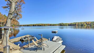 A dock with chairs and a grill, a boat, and a calm lake with trees in the background.