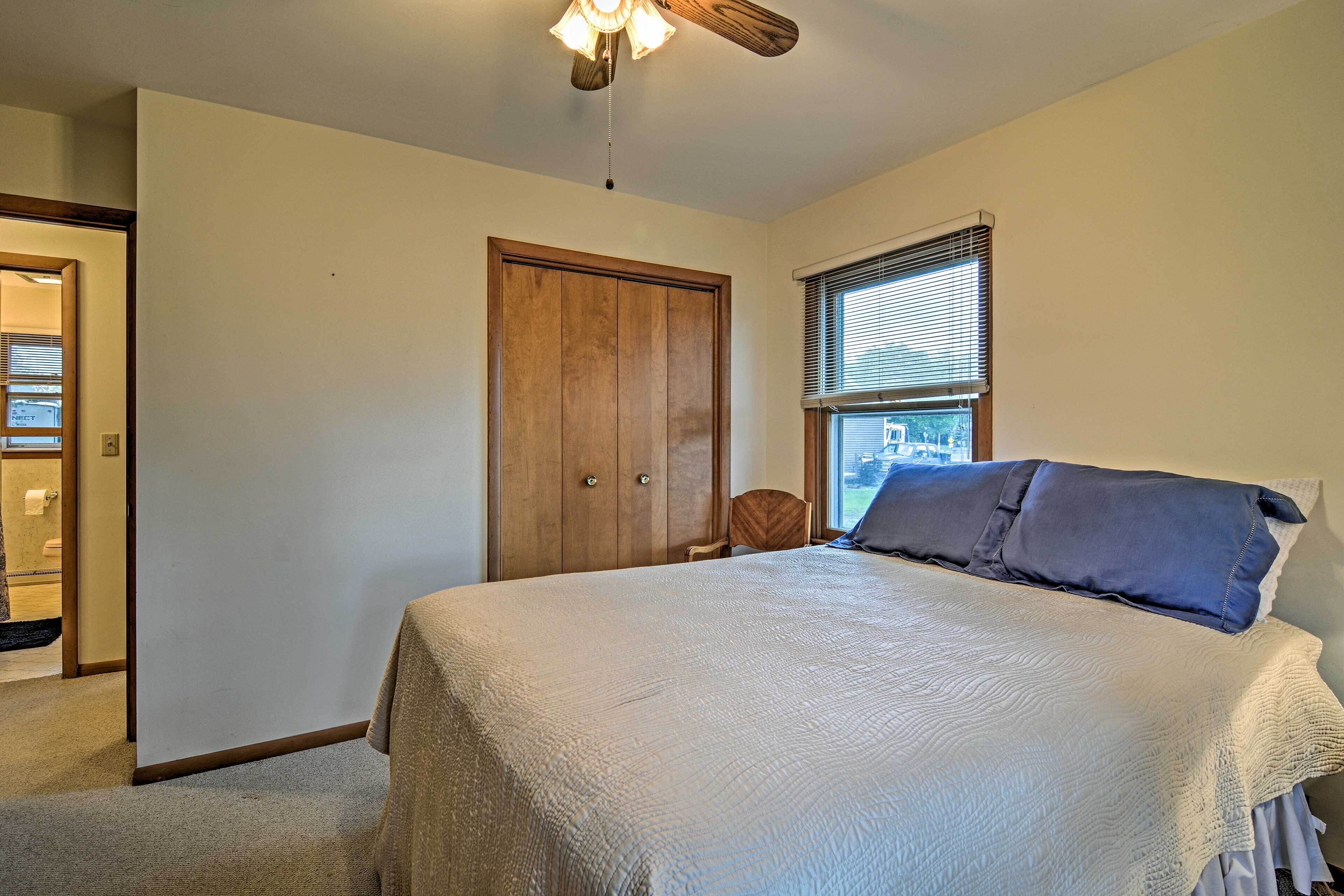Quarto com cama, guarda-roupa de madeira, janela com persianas e ventoinha no teto.