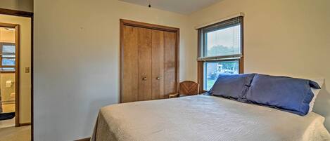 Quarto com cama, guarda-roupa de madeira, janela com persianas e ventoinha no teto.