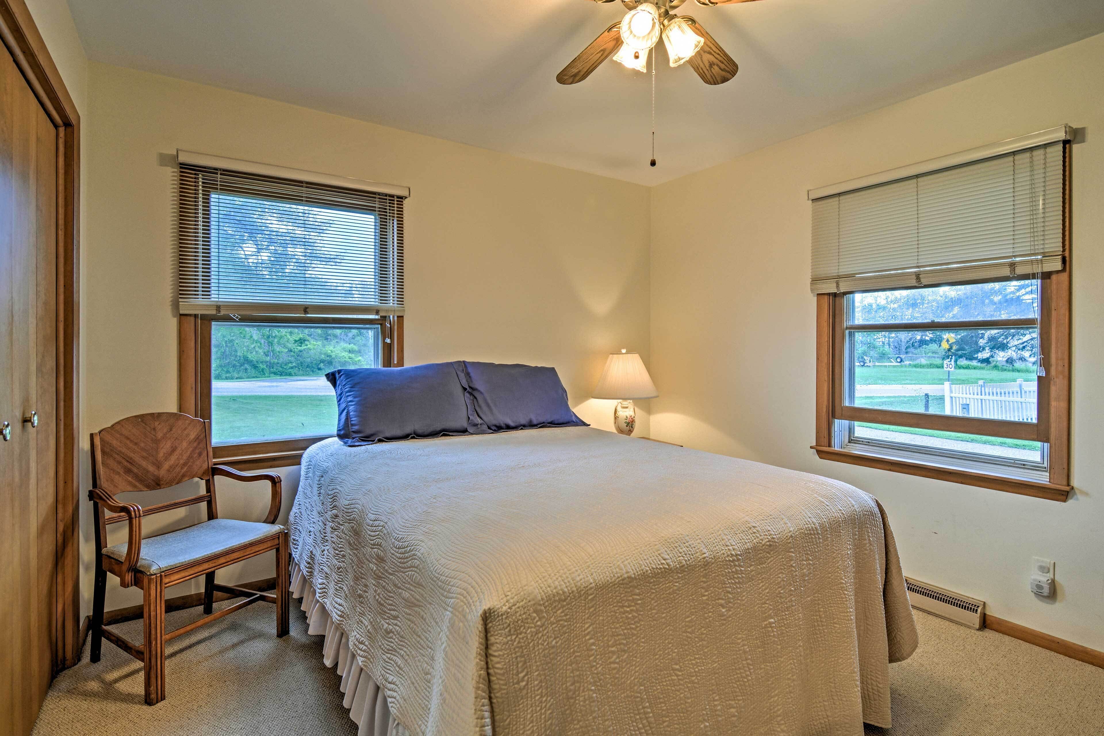A bedroom with a bed, a chair, two windows with blinds, a ceiling fan, and a nightstand with a lamp.