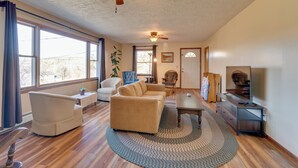 A spacious living room with a large window, a ceiling fan, a sofa, a coffee table, and a television. - Peaceful Horseheads Hideaway w/ Deck & Yard! (Horseheads)