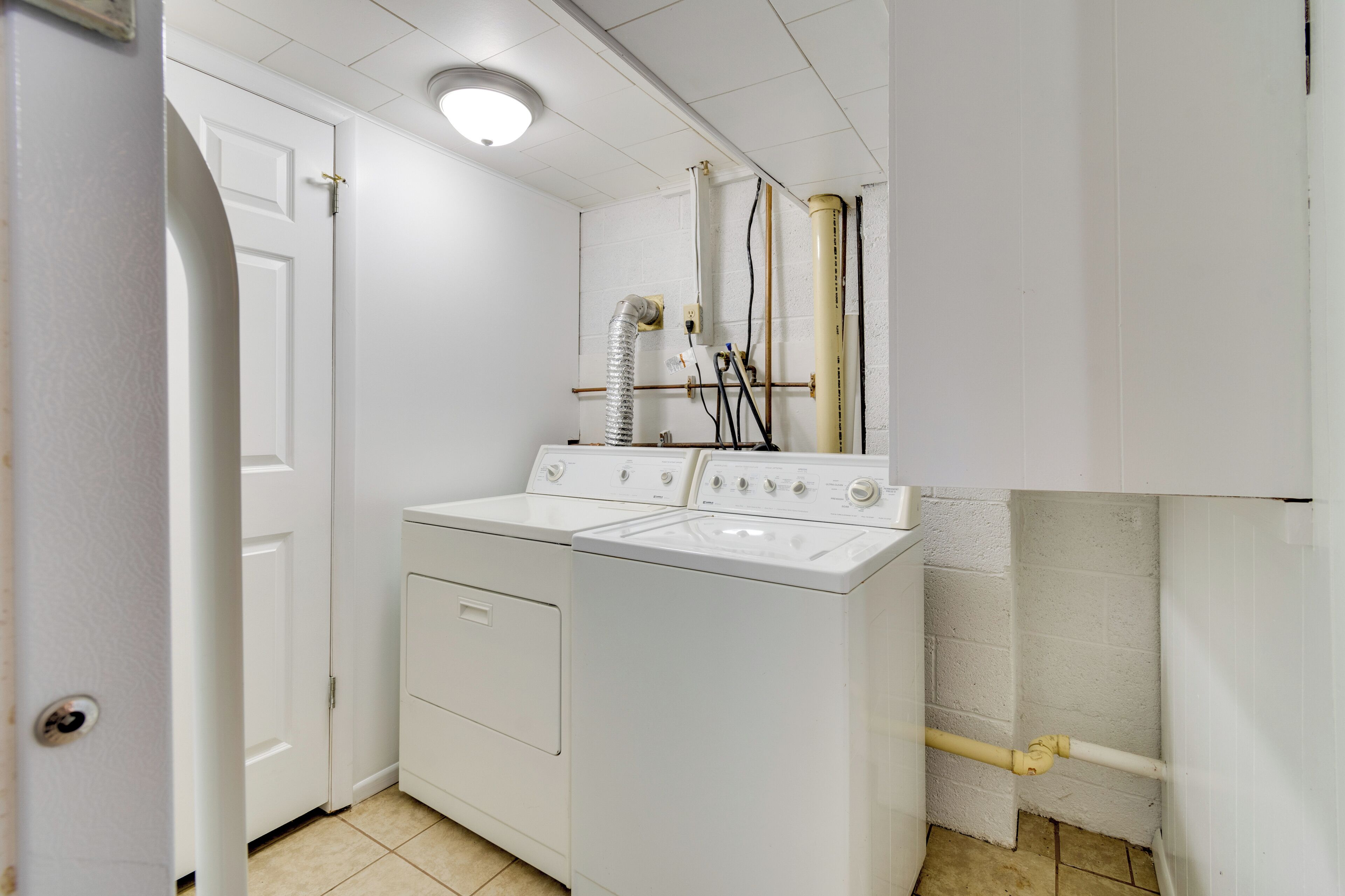 A laundry room with two white washers and dryers, a vent, and a door leading to another room.