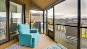 A covered balcony with a blue chair, a small table with a lamp, and a view of a deck and mountains.