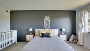 Un dormitorio con una cama grande, dos mesitas de noche, un atrapasueños, una cuna y una ventana con cortinas.