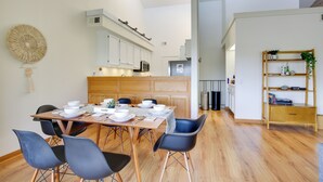 Un comedor moderno con una mesa de madera dispuesta para cuatro personas, rodeada de sillas azules. Se ve una cocina con armarios blancos y un microondas al fondo.