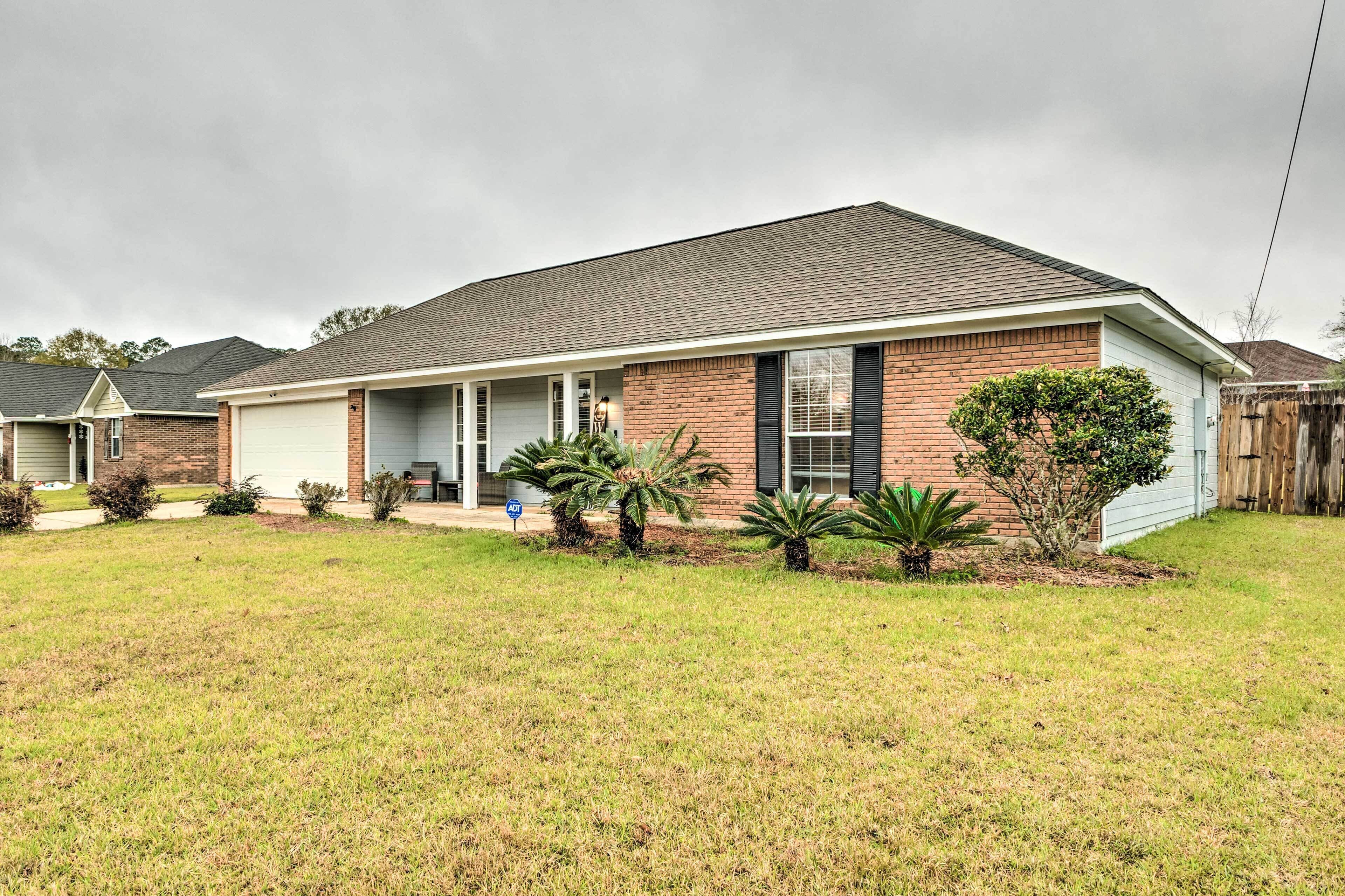 A single-story brick house with a front garage, a covered porch, and a small front yard with various plants.