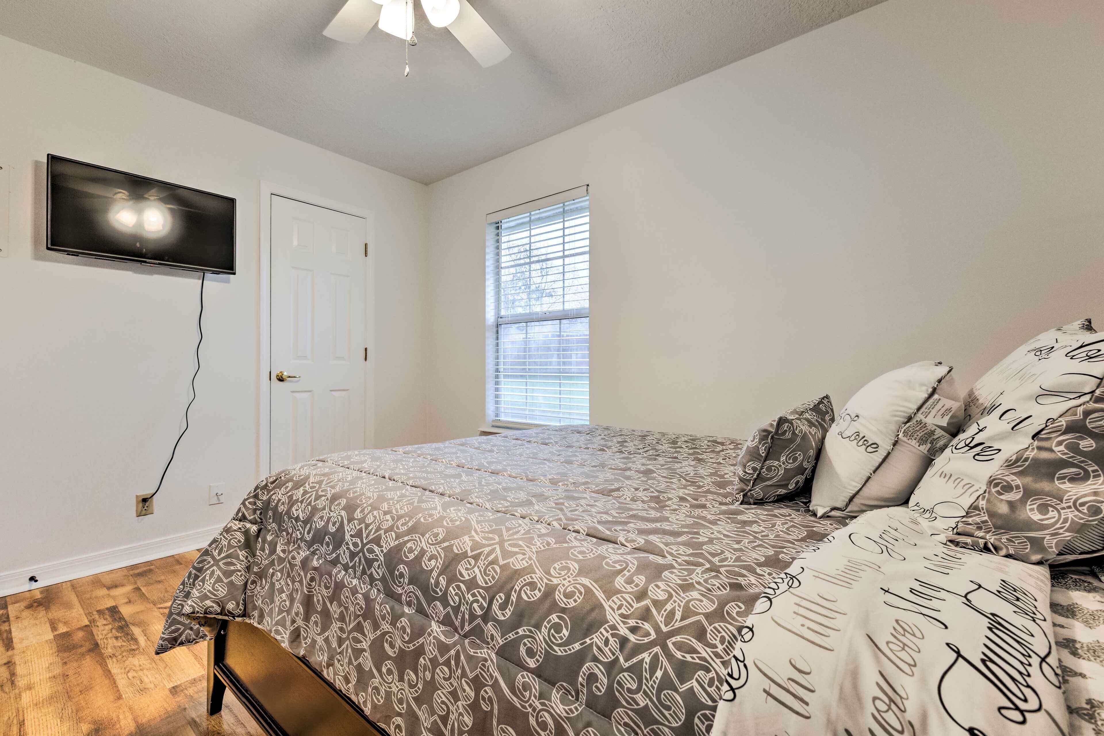A bedroom with a bed, a TV mounted on the wall, a window with blinds, and a door.