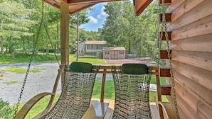 A wooden porch with two wicker chairs and a swing, overlooking a gravel path and a house in a wooded area.