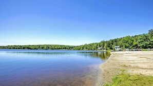 A serene lakeside view with a clear sky, calm water, and a forested shoreline. - Walk to Beach: Lakefront Pocono Escape w/ Fire Pit (Pocono Lake)