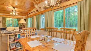 A wooden dining table with chairs, a chandelier, and a view of the forest through large windows. - Secluded Farwell Cabin w/ Fire Pit & Gas Grill! (Farwell)