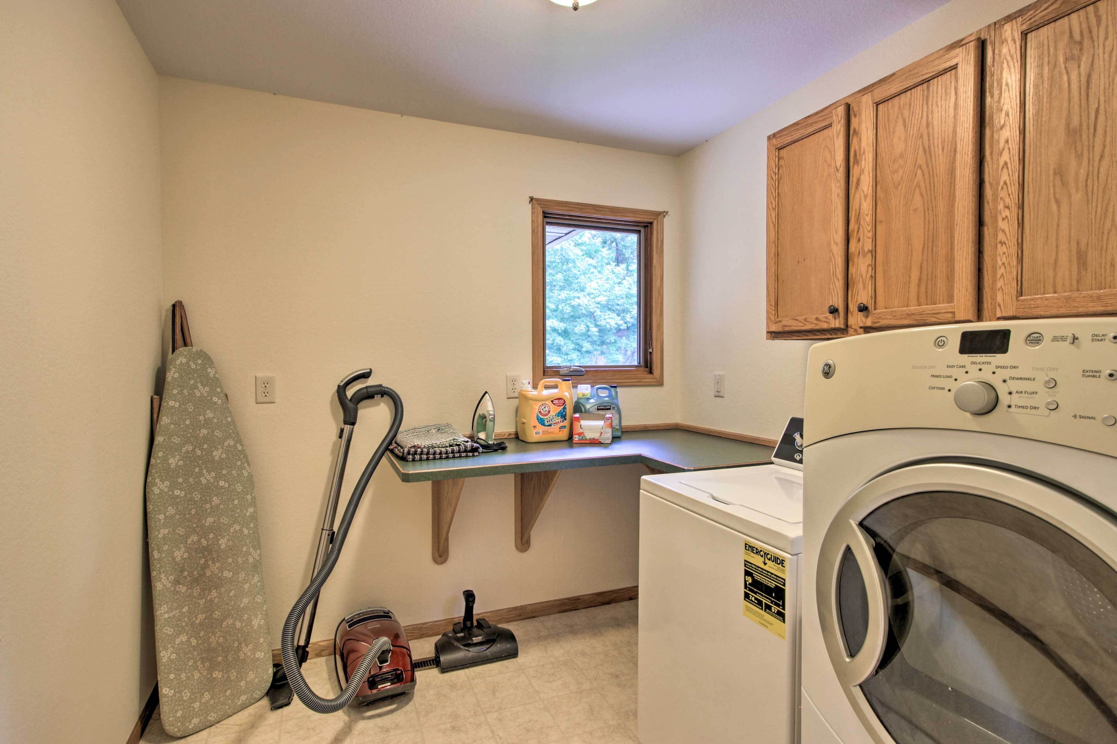 A laundry room with a washing machine, ironing board, vacuum cleaner, and laundry supplies.