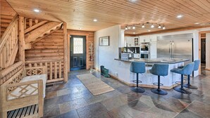 A log cabin interior with a wooden staircase, a kitchen area with a refrigerator, and a bar with stools.