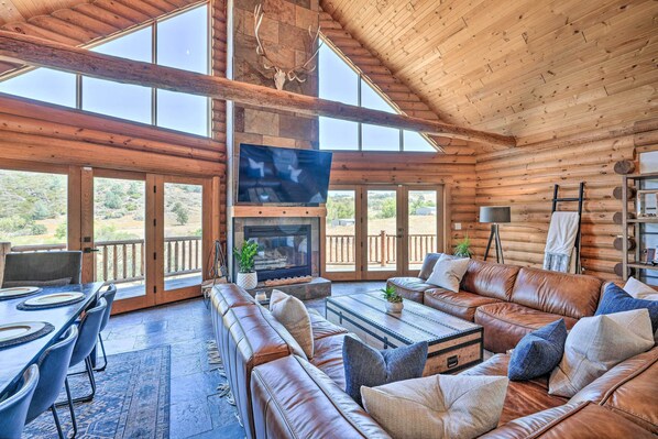 A spacious log cabin with a large fireplace, leather seating, and large windows offering a view of the surrounding landscape.